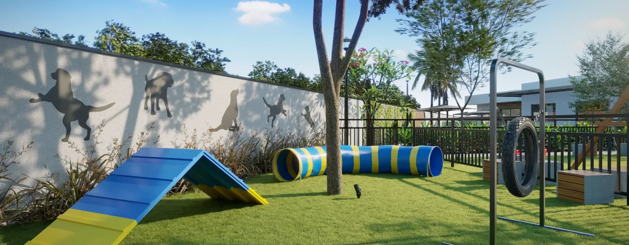 Área pet play do condomínio com rampa azul e amarela, túnel listrado e pneu suspenso para agility sobre gramado, cercada por muro com silhuetas de cães e vegetação.
