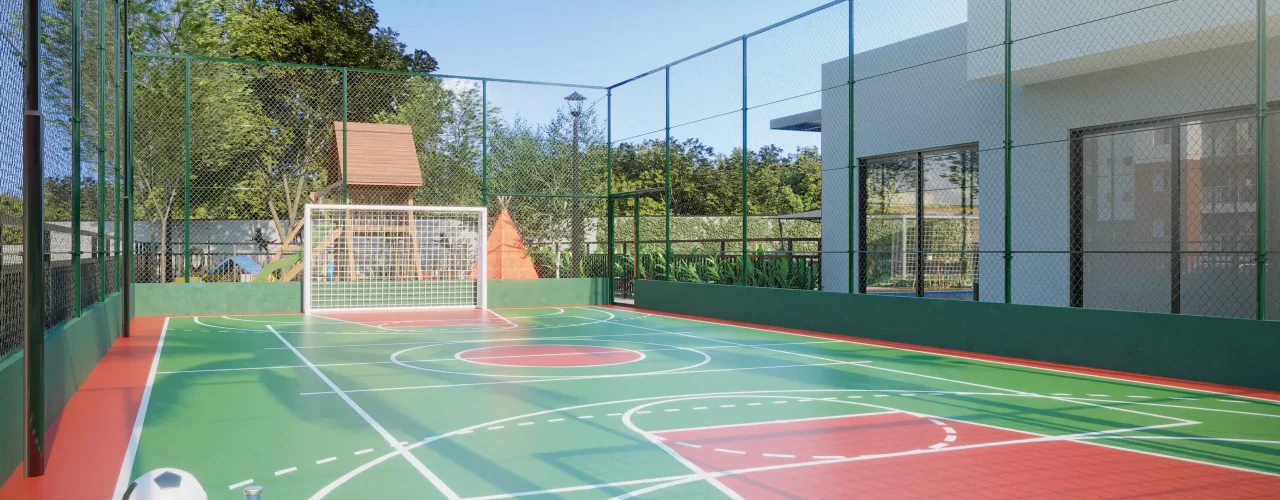 Quadra esportiva do condomínio com piso verde e marcações brancas, bola de futebol, garrafa e toalha no canto, cercada por tela de proteção e gol ao fundo, pronta para prática de esportes ao ar livre