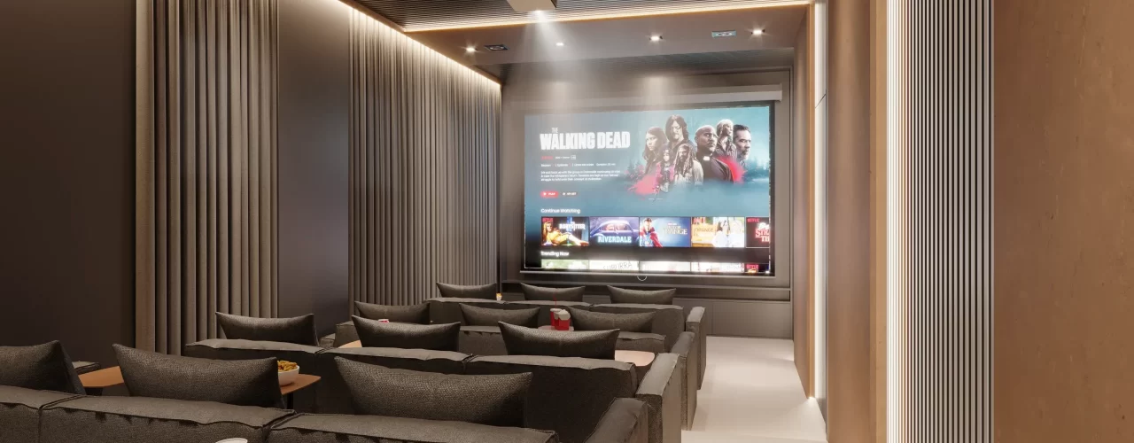 Sala de cinema do condomínio com sofás confortáveis, projetor ligado e tela exibindo catálogo de filmes, acompanhada de mesas de apoio com pipoca e refrigerante.